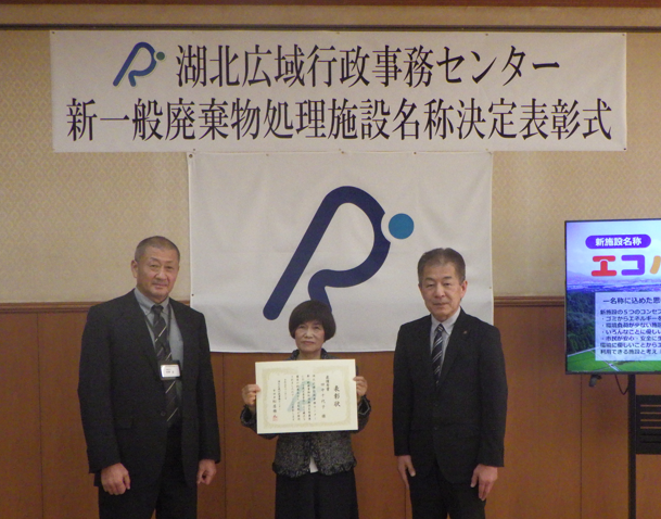 表彰状を手にされている田中さんと秋野事務局長、松井管理者がセンターのシンボルマークの旗の前に並んでいる写真