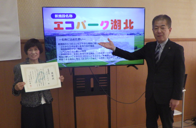 表彰状を手にされている田中さんとモニターに映った「エコパーク湖北」の文字を紹介する松居管理者の写真
